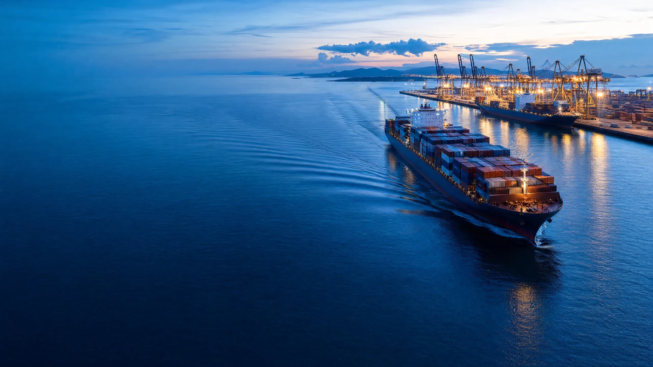 Buque portacontenedores navegando hacia un puerto iluminado a la hora azul